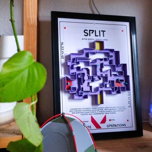 Puede incluir: Obra de arte enmarcada con un mapa 3D púrpura etiquetado como "SPLIT" con texto que indica Alpha Earth, Tokio Japón. El mapa está sobre un fondo blanco con detalles en rojo y amarillo. Una pequeña tienda de campaña está delante.