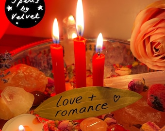 May include: Three lit red taper candles arranged on a silver tray with crystals, dried rosebuds, and a bay leaf inscribed with the text love + romance. A small tea light candle glows nearby, and a large peach-coloured rose sits on the right side.
