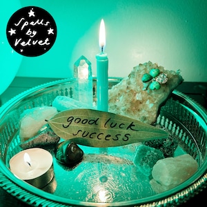 May include: A silver-toned metal tray holds a lit taper candle, a tea light, a clear crystal point, a cluster of crystals with a green clover pin, and a bay leaf inscribed with the words good luck success in black ink under green lighting.