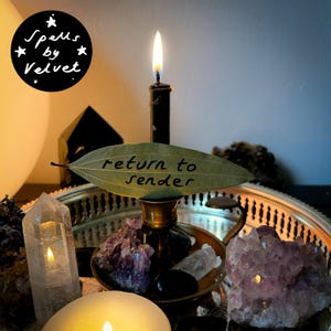 Può includere: Una candela nera in un portacandele di ottone con una foglia di alloro che riporta la scritta return to sender. La candela è circondata da grappoli di ametista, punte di quarzo trasparente e un vassoio argentato. Un logo indica Spells by Velvet.