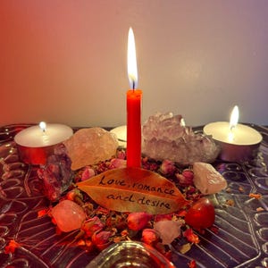 May include: A burning red candle stands central, encircled by crystals, dried rose petals, and two lit tea lights. A leaf inscribed with "Love, romance and desire" is in the foreground. The arrangement is on a decorative silver tray.
