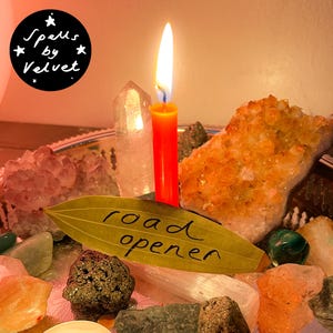 Puede incluir: Una vela naranja encendida se encuentra entre varios cristales, incluyendo un gran racimo naranja, una punta transparente y piedras más pequeñas. Una hoja seca con el texto escrito a mano "road opener" descansa delante. Un logotipo circular negro dice "Spells by Velvet".