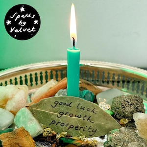 Peut inclure: Une bougie sarcelle allumée est posée sur un plateau argenté entourée de cristaux, de pierres et d'une feuille de laurier portant le texte manuscrit good luck growth prosperity. Un logo noir affiche le texte Spells by Velvet.