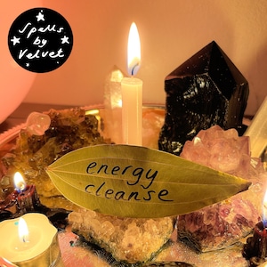 May include: A dried bay leaf with the words energy cleanse written in black ink, positioned in front of a lit white taper candle, surrounded by various crystals including amethyst and black obsidian, with a Spells by Velvet logo in the corner.
