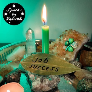 May include: A green candle with a lit flame stands behind a dried bay leaf inscribed with the words Job Success. The candle is surrounded by various crystals, including a large citrine cluster with a green four-leaf clover pin attached.