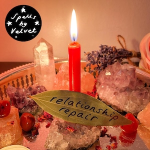Puede incluir: Una vela roja con llama encendida sobre una bandeja plateada rodeada de cristales de amatista y cuarzo transparente, lavanda seca y piedras rojas. Una hoja de laurel con el texto escrito a mano relationship repair descansa delante.
