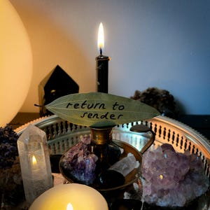 May include: A burning black candle in a brass candlestick holder on a silver tray, with crystals and a leaf inscribed with "return to sender". The image has a soft, mystical lighting, creating a magical atmosphere.