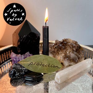 May include: A silver tray holds a black candle with a lit flame, a black obsidian point, a smoky quartz cluster, a selenite wand, a piece of amethyst, two black stones, and a bay leaf with the word protection written on it in black ink.