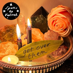 May include: A silver-toned metal tray holds two lit black taper candles, a white tea light, various raw crystals in shades of orange and clear, a dark obsidian point, and a peach-coloured rose. A green leaf with the text get over them is present.