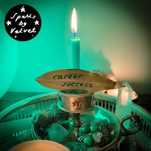 Puede incluir: Una vela azul claro arde en un candelabro de metal plateado con recortes de estrellas. Una hoja seca con las palabras career success escritas en tinta negra se coloca frente a la vela. La escena está iluminada por una luz verde brillante.