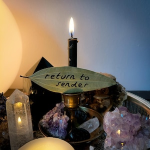 Pode incluir: Uma vela preta acesa em um castiçal de latão com uma folha verde que diz "return to sender". A vela está em uma bandeja de prata cercada por cristais e outros itens espirituais. Uma grande esfera branca ao fundo.