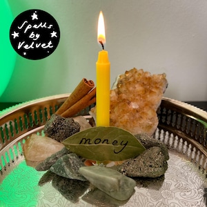 May include: A lit yellow taper candle stands on a silver tray surrounded by a cluster of citrine crystals, green stones, two cinnamon sticks, and a single bay leaf with the word money written in black ink. A black circular logo reads Spells by Velvet.