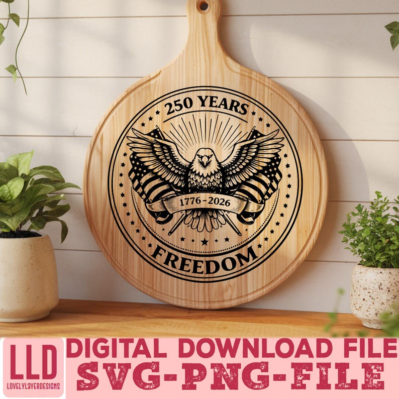 May include: A round wooden cutting board with a handle, featuring a detailed black graphic of an eagle with outstretched wings, American flags, and the text "250 YEARS 1776-2026 FREEDOM". The board is light brown.