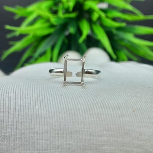 May include: A silver ring with a rectangular setting, designed to hold a gemstone. The ring is set against a white fabric background, with green foliage in the background.
