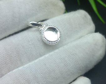 May include: A small, silver pendant with a circular setting and a textured rope-like border. The pendant has a loop for a chain and is displayed on a white fabric surface.