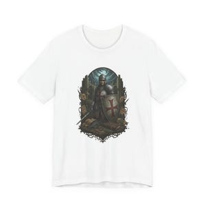 May include: White t-shirt with a detailed illustration of a medieval knight in full armour, holding a shield with a red cross. The knight stands before a backdrop of a castle and fallen swords. The design is surrounded by a dark, ornate border.
