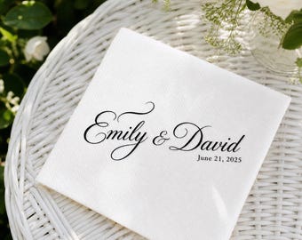 May include: A white linen napkin with the names "Emily & David" in elegant black script, along with the date "June 21, 2025". The napkin is placed on a white wicker surface, with greenery and white flowers in the background.