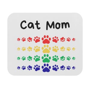 Könnte beinhalten: Weißes quadratisches Mauspad mit dem Text "Cat Mom" in Schwarz. Darunter befinden sich Reihen farbiger Pfotenabdrücke in Rot, Gelb, Grün und Blau. Ein lustiges Accessoire für Katzenliebhaber.