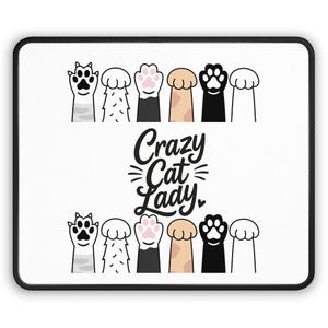 Puede incluir: Alfombrilla de ratón rectangular blanca con borde negro. La alfombrilla presenta un diseño de patas de gato en varios colores y el texto "Crazy Cat Lady" en una fuente de escritura.