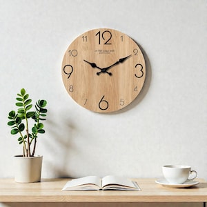 May include: A circular wooden wall clock with black numerals and hands. The clock face has a light wood grain pattern. The text "est 1765 vintage studio QUARTZ CLOCK" is printed above the centre. A small potted plant, an open book, and a teacup are on a wooden surface.