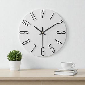 May include: A white, round wall clock with black numerals and hands. The clock has a minimalist design, with the numbers in a simple, modern font. The clock is mounted on a white wall. A small potted plant and a cup of coffee are on a wooden surface.