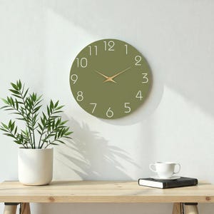 Puede incluir: Reloj de pared redondo verde oliva con números blancos y manecillas doradas. El reloj está montado en una pared blanca. Una planta en maceta y una taza de café están sobre una mesa de madera.