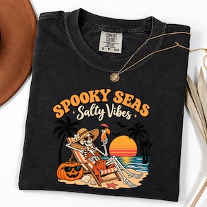 May include: A black t-shirt featuring a graphic of a skeleton wearing a sun hat and sunglasses, lounging in a beach chair with a drink. The text reads "SPOOKY SEAS Salty Vibes" above a sunset, palm trees, and a jack-o'-lantern on the sandy beach.