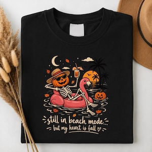 May include: A black t-shirt featuring a graphic of a skeleton with a pumpkin head wearing sunglasses and a straw hat, lounging on a pink flamingo pool float in water. The design includes palm trees, a crescent moon, and the text still in beach mode... but my heart is fall.