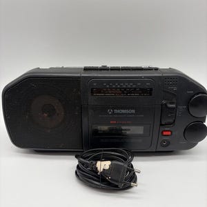 Könnte beinhalten: Ein schwarzes, tragbares Thomson AM/FM-Radio-Kassettendeck mit Lautsprechergitter auf der linken Seite, zentralem Kassettendeck, Radiofrequenzanzeige und zwei runden Drehreglern auf der rechten Seite. Ein schwarzes Netzkabel liegt davor aufgerollt.