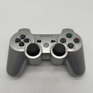 May include: A silver Sony PlayStation game controller featuring two black analog sticks, a directional pad, four action buttons with geometric symbols, a central PlayStation logo button, and Select and Start buttons labeled in black text.