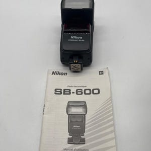 Peut inclure: Un flash électronique Nikon Speedlight SB-600 noir avec une tête rectangulaire et un pied de montage en métal, accompagné d'un manuel d'instruction blanc portant le texte Nikon Flash électronique SB-600 Manuel d'utilisation.