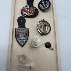 May include: A collection of vintage military and service insignia including a black leather shoulder board with a shield, a circular black and silver torch emblem, a shield-shaped Lyon fire department badge, a silver horn pin, and two small pins in plastic bags.
