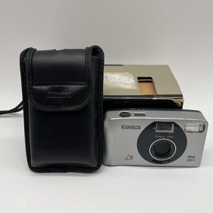 May include: A silver Konica Mini compact film camera with a black lens housing, flash, and viewfinder, positioned next to a black leather carrying case embossed with the Konica logo and a cardboard box with a rectangular cutout on the top surface.