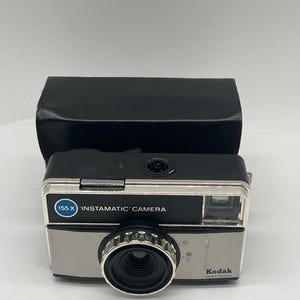 May include: A black and silver Kodak 155 X Instamatic camera with a rectangular body, a central circular lens, and a black protective case positioned behind the camera body. The front panel features the text 155 X INSTAMATIC CAMERA and Kodak.