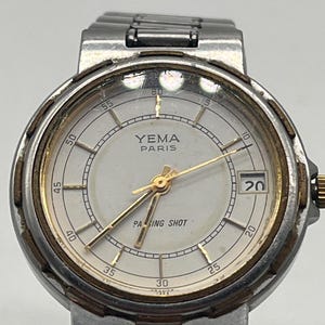 Peut inclure: Une montre-bracelet Yema Paris Passing Shot avec un bracelet en métal argenté et un cadran rond blanc. Le cadran présente des aiguilles dorées, des index dorés, un guichet de date à trois heures et une lunette dorée avec des accents marron.