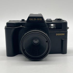 Può includere: Una fotocamera nera con obiettivo centrale, caratterizzata dalla scritta QLS-99 stampata in oro sulla parte superiore e 2000N stampata in oro sul lato anteriore destro. Il dispositivo ha una finitura nero opaco, un mirino e una ghiera.