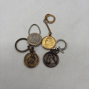 May include: Four circular metal keychains featuring religious imagery. One gold-toned keychain depicts a profile of a figure, while the others show embossed scenes of figures in silver and bronze tones. Each keychain includes a metal ring attachment.