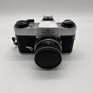 Op de afbeelding: Een zilveren en zwarte IFBAFLEX 35mm filmcamera met een centrale lens met een zwarte dop, een pentaprisma zoekerbehuizing en diverse handmatige bedieningsknoppen op de bovenplaat. De camerabehuizing is bekleed met gestructureerd zwart materiaal.