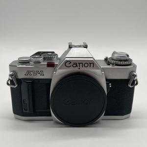 Könnte beinhalten: Eine silber-schwarze Canon AV-1 35mm Filmkameragehäuse mit schwarzem Objektivdeckel. Die Kamera verfügt über eine silberne Deckplatte mit verschiedenen Einstellrädern, einen schwarzen strukturierten Griff, einen Blitzschuh und Metallösen.