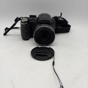 Peut inclure: Un appareil photo numérique noir Fujifilm avec un objectif Super EBC Fujinon doté d'un zoom 26x. L'appareil comprend un cache d'objectif noir amovible avec une cordelette et une sangle de cou noire, présenté de face.