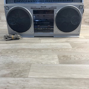 Może przedstawiać: Srebrny radiomagnetofon Philips z odtwarzaczem kaset, dwoma dużymi okrągłymi czarnymi głośnikami oraz wyświetlaczem tunera radiowego z oznaczeniami częstotliwości. Urządzenie posiada zwinięty przewód zasilający oraz napis Philips i model D8434.