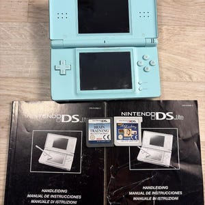 Könnte beinhalten: Eine hellblaue Nintendo DS Lite Handheld-Spielkonsole mit zwei Bildschirmen und Steuertasten, begleitet von zwei Spielkassetten mit der Aufschrift Brain Training und Art Academy sowie zwei schwarzen Bedienungsanleitungen.