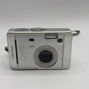 Puede incluir: Una cámara digital Pentax Optio plateada con acabado de metal cepillado. El frente presenta un objetivo con la etiqueta 3x OPT. ZOOM, 5.8mm-17.4mm y 4.1 Megapixels. La palabra Optio está impresa verticalmente a la izquierda y un flash arriba.