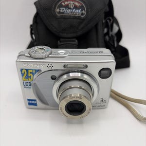 Op de afbeelding: Een zilveren Sony Cyber-shot digitale camera met een Carl Zeiss Vario-Tessar lens, voorzien van een 2,5 inch LCD-monitor label, 3x optische zoom en 5.1 megapixels tekst. De camera staat voor een zwarte Tamrac Digital Series tas.