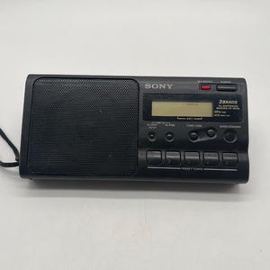 Könnte beinhalten: Ein schwarzes tragbares Sony-Radio mit einem rechteckigen Lautsprechergitter auf der linken Seite und einem digitalen Display auf der rechten Seite. Das Gerät verfügt über einen Ein-/Ausschalter, eine Tastensperre, Regler und fünf Stationstasten.
