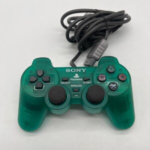 Könnte beinhalten: Ein durchscheinender smaragdgrüner Sony PlayStation-Gamecontroller mit schwarzen Analogsticks, einem Steuerkreuz, vier Aktionstasten mit Symbolen und zentralen Tasten mit der Aufschrift SONY, PlayStation, SELECT, START und ANALOG, inklusive Kabel.