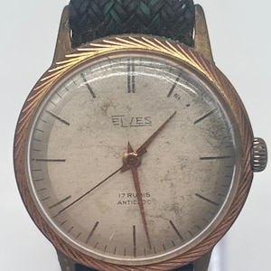 Op de afbeelding: Een vintage polshorloge met een ronde goudkleurige kast, een getextureerde rand en een verweerde gebroken witte wijzerplaat. Op de wijzerplaat staat de tekst ELVES, 17 RUBIS en ANTICLOC. Het horloge heeft dunne gouden wijzers en een donkergroene gevlochten stoffen band.