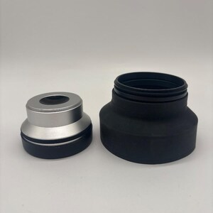 Può includere: Un set di adattatori per obiettivi fotografici in due pezzi composto da un componente metallico più piccolo color argento e nero con un'apertura circolare e un componente cilindrico nero più largo con una sezione superiore filettata.