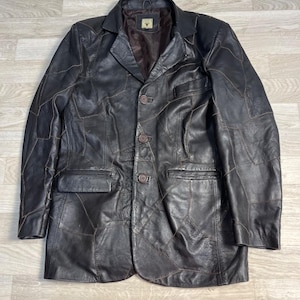 Pode incluir: Um blazer de couro castanho escuro construído a partir de múltiplos painéis costurados. O casaco apresenta uma lapela entalhada, três botões frontais, dois bolsos laterais com aba e um bolso no peito. O interior é forrado com tecido.