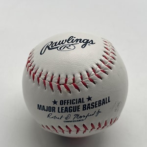 Peut inclure: Une balle de baseball en cuir blanc avec des coutures rouges, arborant le logo Rawlings en bleu foncé. La balle porte l'inscription Official Major League Baseball et la signature du commissaire Robert D. Manfred Jr. en encre bleu foncé.